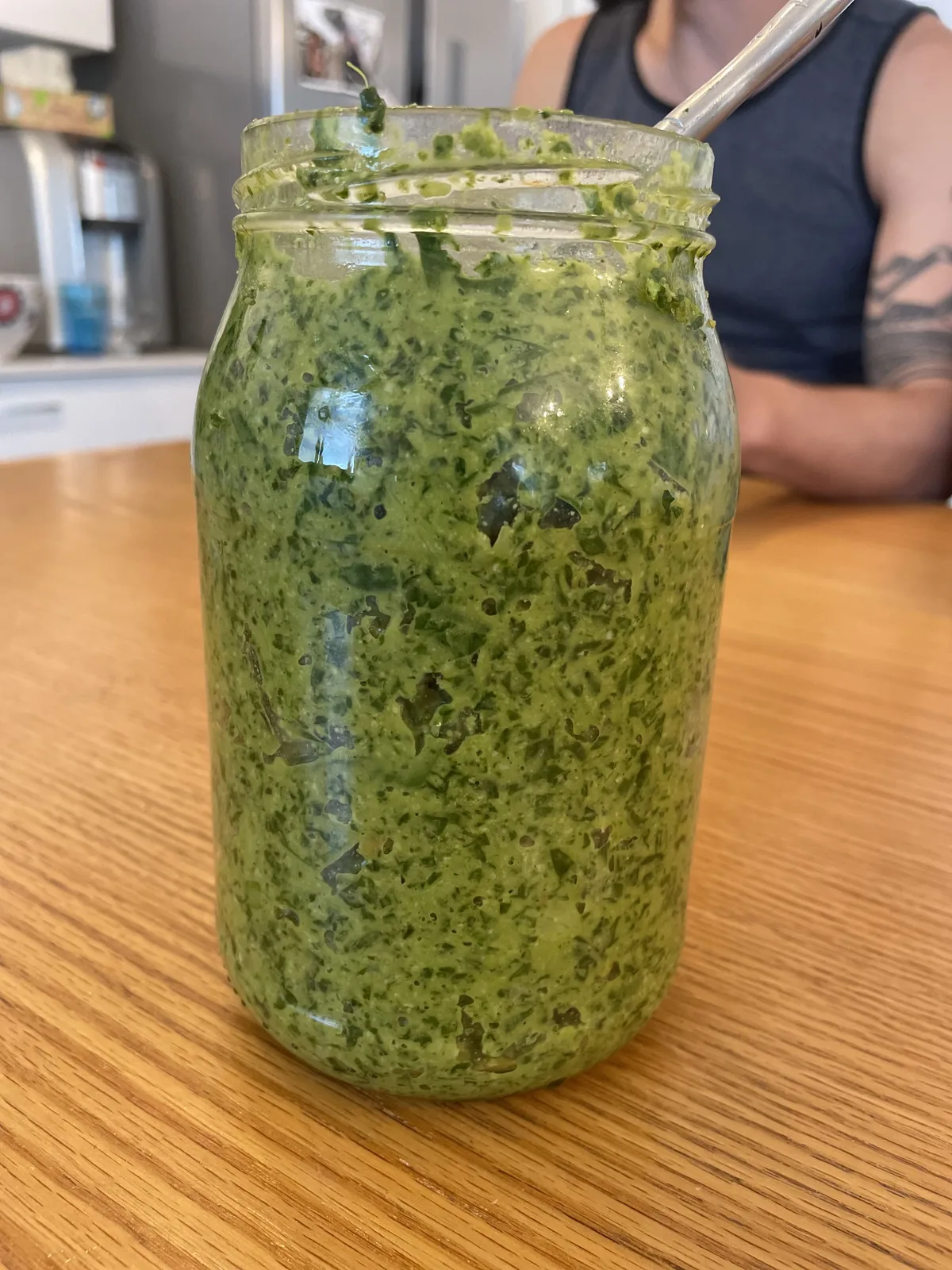 A wide-mouth glass jar of deep-green ayurvedic pesto with a thin layer of olive oil on top, surrounded by fresh basil, cilantro, parsley, garlic cloves, and a small bowl of pine nuts