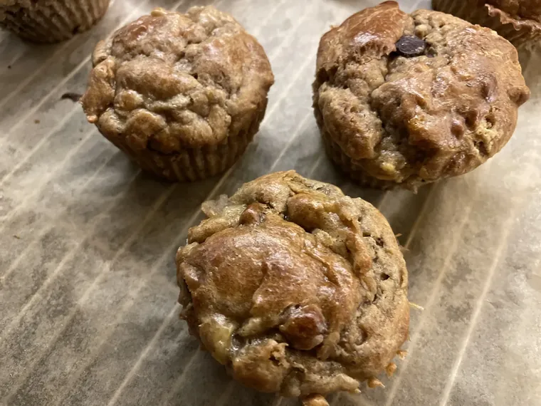 A dozen golden flourless muffins on a wire rack, studded with blueberries and slices of ripe banana, one cut open to show the soft cashew-butter crumb