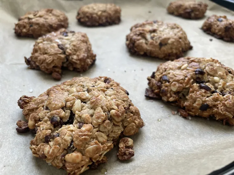 A dozen golden flourless tahini cookies cooling on parchment paper, studded with dark cocoa nibs and pine nuts, with flaky Himalayan salt on top