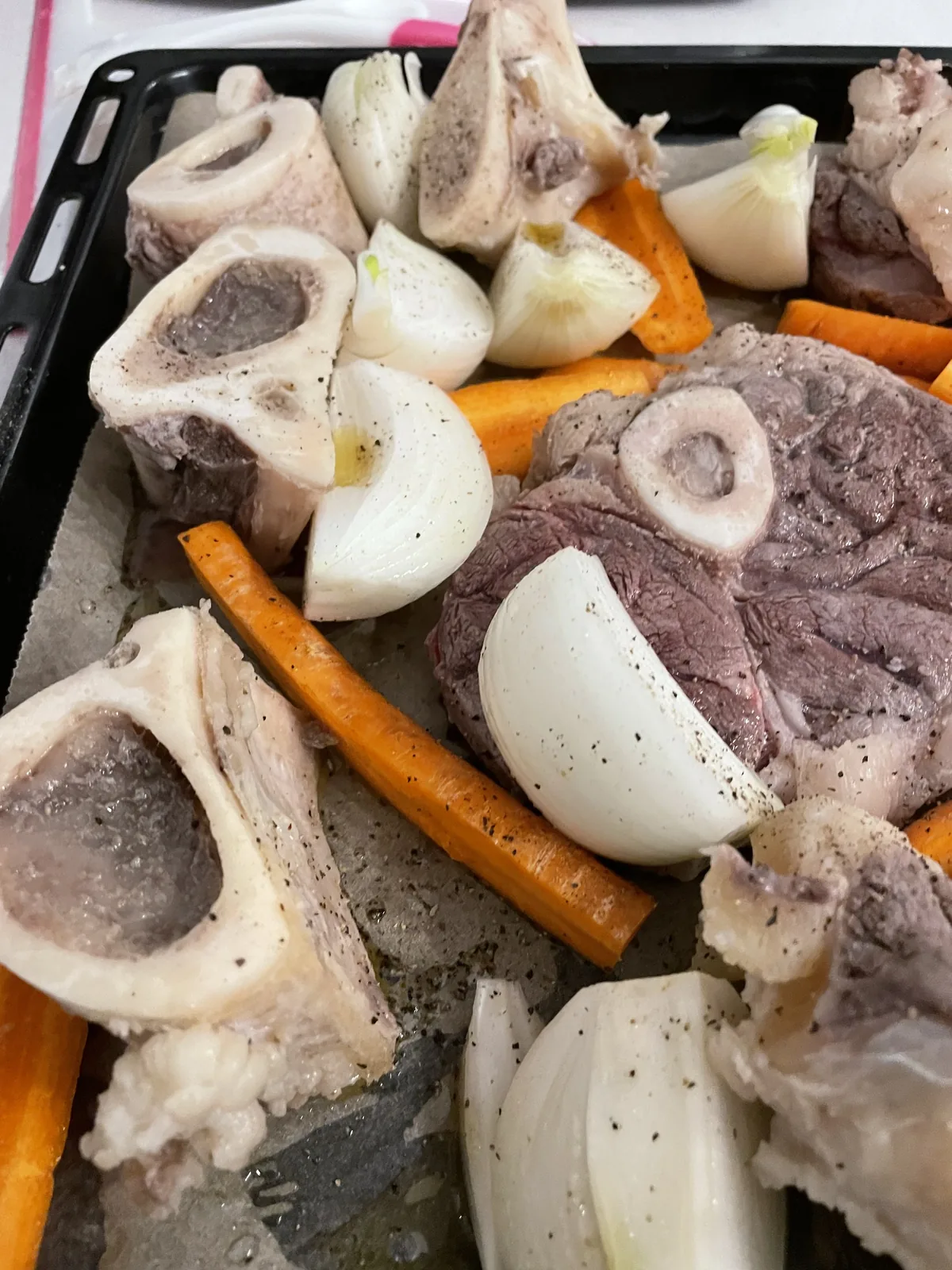 Pre-roast sheet pan showing fresh beef marrow bones, cross-cut ossobuco shank, quartered onions, whole carrots, and freshly ground black pepper — ready for the oven before the long simmer