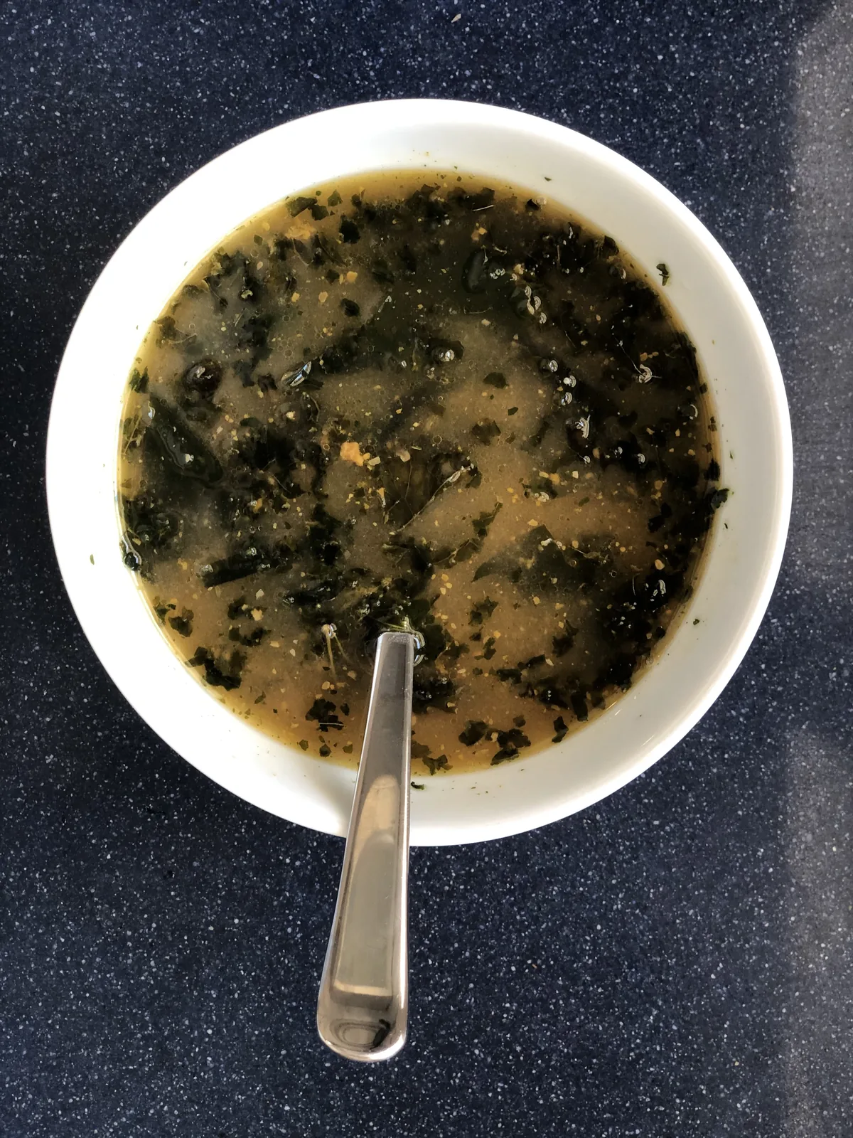 A white bowl of finished instant bone broth soup with kale flakes and seaweed floating on the surface, spoon ready beside it