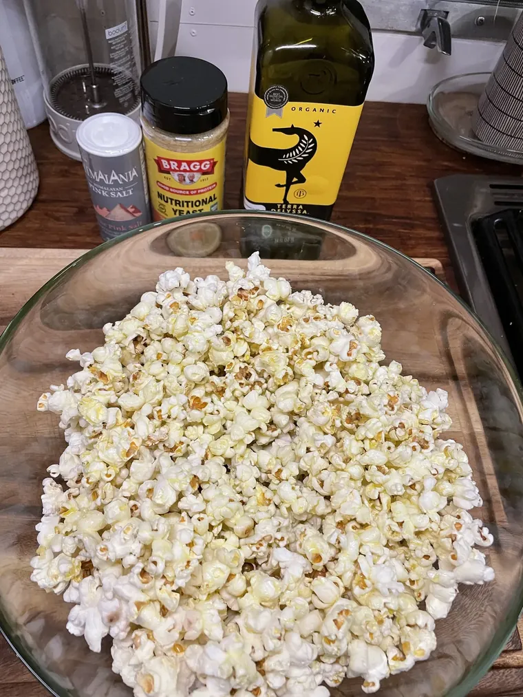 A wooden bowl of freshly popped popcorn drizzled with olive oil, dusted golden with nutritional yeast and flecked with pink Himalayan salt