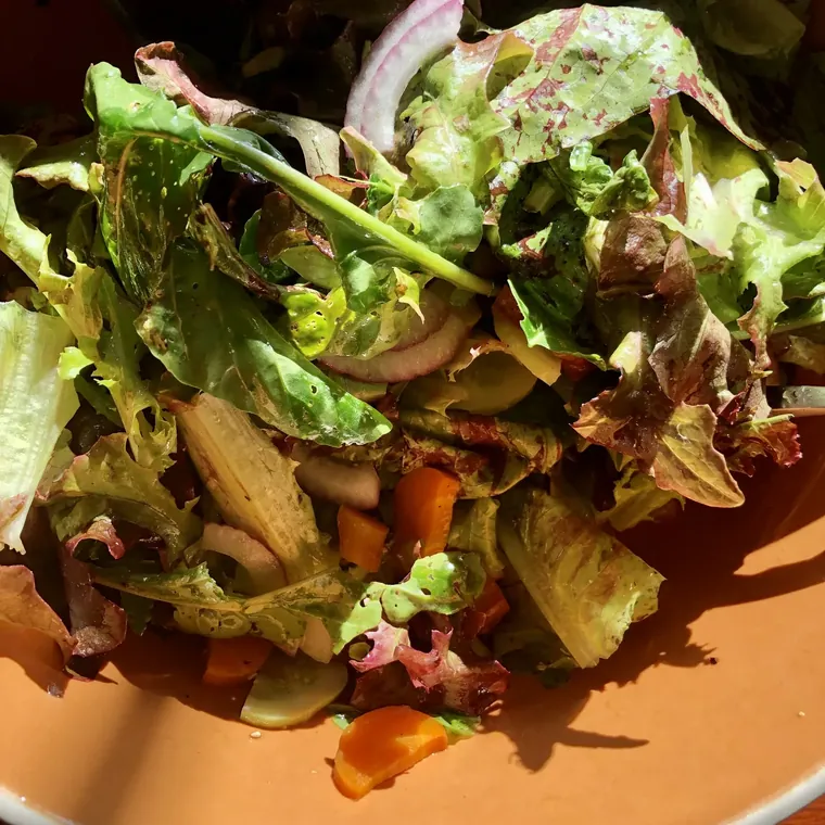 Large wooden bowl of butter lettuce and arugula with thin red onion rings, dry-roasted pumpkin seeds, and a glossy creamy tahini dressing — set on a long potluck table